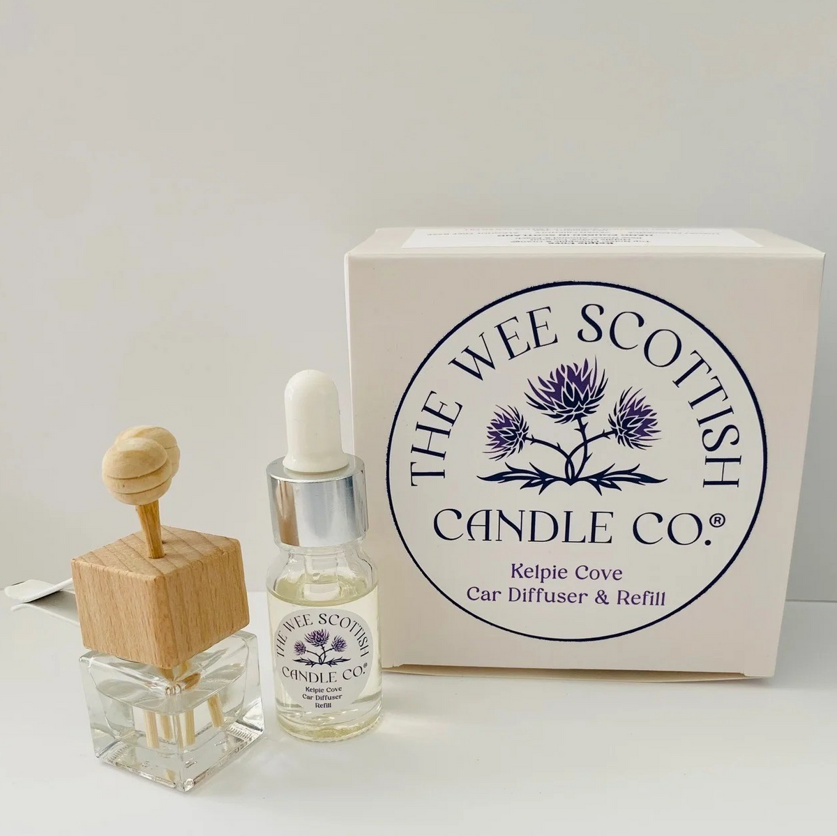 The Wee Scottish Candle Co. Kelpie Cove car diffuser and refill set, featuring a small glass bottle with a dropper, a square glass car diffuser with a wooden honey pot top, and the white product packaging box.