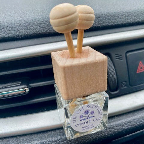 A car air freshener diffuser with a square light wood block top and two round wooden reeds, inserted into a clear glass bottle with a 'The Wee Scottish Candle Co.' label, resting on a car dashboard.